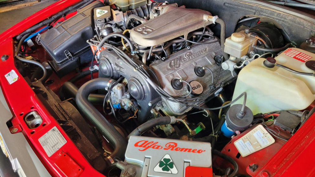 Nahaufnahme des Motorraums eines roten Alfa Romeo. Zu sehen sind verschiedene Motorkomponenten, Schläuche und Behälter. Auf einer Abdeckung ist das Alfa Romeo-Emblem deutlich zu erkennen.